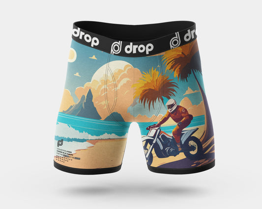 A pair of men's boxer briefs with a colorful retro beach scene print, featuring a rider on a dirt bike, palm trees, mountains, and a large sun. The waistband is black with the brand name 'drop' repeated in white.